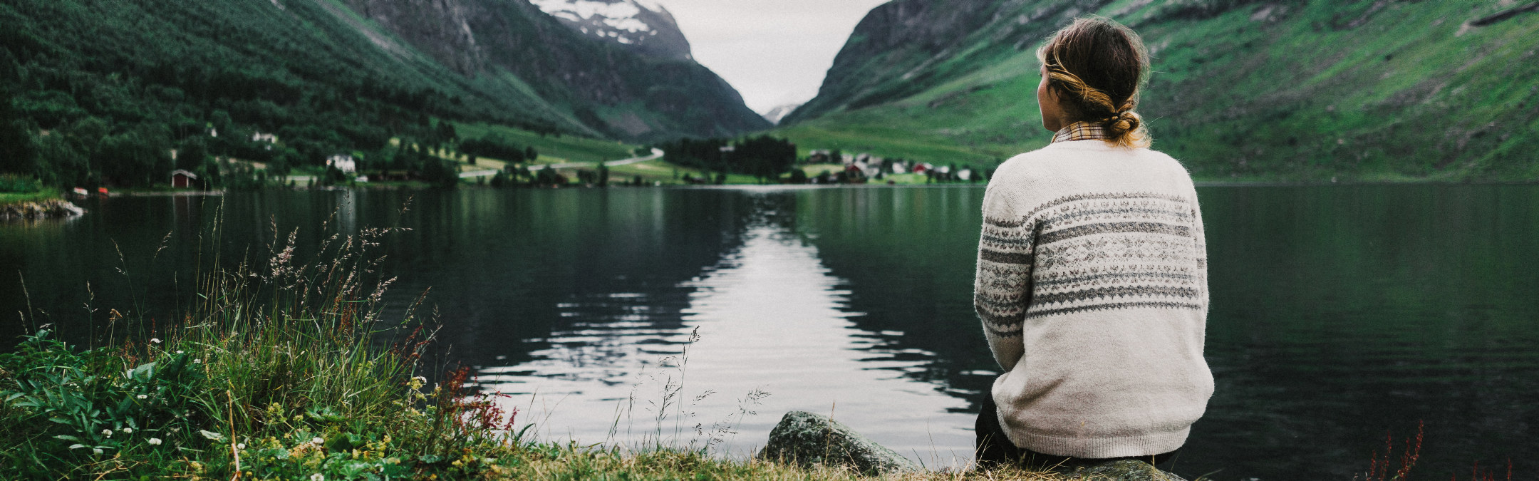 En unge dame i strikket genser sitter og ser utover en innsjø i grønne omgivelser. 